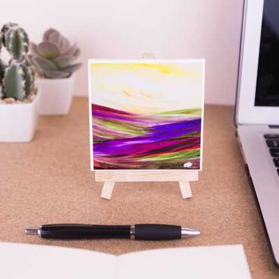 Mini easel holding ceramic coaster of a sheep looking ahead at the expanse of the purple moorland with a golden sky