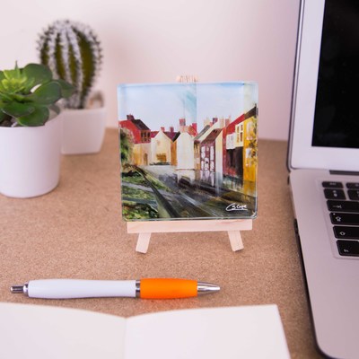 A glass coaster of the painting 'Greatham in the rain' is sitting on a mini easel
